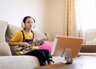 Niño consciente en auriculares con los ojos cerrados y saxofón en el sofá grabación de vídeo en el teléfono celular en casa - foto de stock