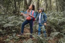 Ethnisches Mädchen mit Papierleitfaden schaut mit Fernglas zwischen Farnpflanzen im Sommerwald gegen Bruder weg — Stockfoto