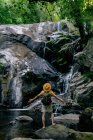 Visão traseira do caminhante masculino irreconhecível em pé em pedra e admirando a cachoeira na floresta com os braços abertos — Fotografia de Stock