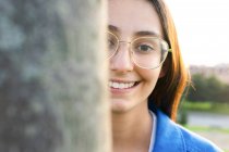 Fröhliche Frau mit Brille guckt aus Baumstamm im Stadtpark und blickt in Kamera — Stockfoto