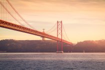 Знаменитый мост 25 de Abril, пересекающий реку Тагус и соединяющий Лиссабон и Алмаду возле святилища Христа Царя, против облачного закатного неба в Португалии — стоковое фото