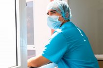 Vista lateral del médico masculino cansado en máscara y uniforme de pie cerca de la ventana en la clínica y teniendo descanso - foto de stock