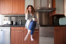 Geschäftsfrau mit lockigem Haar sitzt in der Küche und nimmt einen Aufguss, während sie ihr Smartphone benutzt und zu Hause arbeitet — Stockfoto