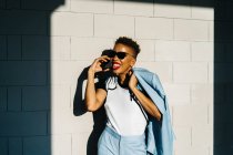 Стильная взрослая афроамериканка с современной стрижкой и курткой, разговаривающая по мобильному телефону со стеной из черепицы с тенью под солнцем — стоковое фото