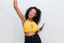Mujer afroamericana feliz en auriculares con el baile del teléfono celular mientras escucha la canción en el fondo de luz - foto de stock
