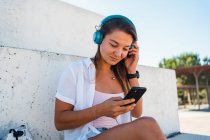 Joven mujer alegre positiva navegando en el teléfono móvil escuchando música en los auriculares en el día soleado en verano en la ciudad - foto de stock