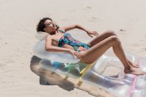 Вид сбоку на счастливую женщину в купальнике лежащую на надувном матрасе на песчаном берегу моря и загорающую в солнечный день во время летних каникул — стоковое фото