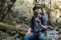 Durstiger männlicher Wanderer mit Rucksack trinkt Kumpel aus Tasse mit Stroh, während er auf Felsen in der Nähe des Wasserfalls im Wald sitzt und in der Pause wegschaut — Stockfoto