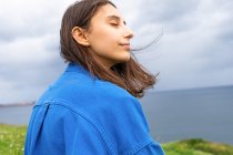 Side view of delighted female with eyes closed sitting on green hill and enjoying view of sea on cloudy day — Stock Photo