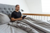 Ruhige Männchen sitzen auf dem Bett unter einer Decke und verwenden Tablette nach dem Aufwachen im Schlafzimmer morgens zu Hause — Stockfoto