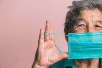 Sorridente invecchiato bocca di copertura femminile con blu maschera medica protettiva da coronavirus mentre guardando la fotocamera su sfondo rosa in studio — Foto stock