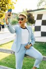 Content mature African American female in modern sunglasses taking selfie on cellphone in park in back lit — Stock Photo
