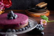 Délicieux gâteau mousse myrtille avec crème pourpre décorée de baies fraîches servi sur support en verre sur table sombre avec des fleurs — Photo de stock
