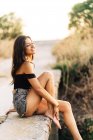Seitenansicht der unbeschwerten barfüßigen jungen hispanischen Frau in schwarzem Top und grauer Jeanshose, die an einem Sommertag in der Natur auf einer Steinbrücke sitzt — Stockfoto