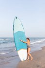 Surfista femenina de pie con tabla SUP azul en la playa de arena en verano y mirando hacia otro lado - foto de stock