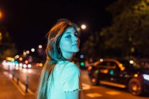 Vista lateral de la encantadora joven hembra con el pelo largo mirando a la cámara contra la carretera en la ciudad de la noche - foto de stock