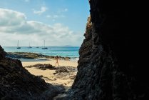 Женщина-туристка, стоящая возле пенных морских волн на мокром песчаном пляже против скалистой скалы и облачного голубого неба во время летних каникул в Фуэртевентуре, Испания — стоковое фото
