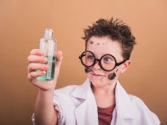 Поразительный мальчик в очках и лабораторном халате с грязным лицом и жидкостью в бутылке после химического эксперимента — стоковое фото