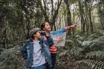 Menina étnica com guia de papel olhando para longe contra o irmão com binóculos entre plantas de samambaia em florestas de verão — Fotografia de Stock