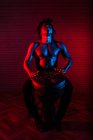 Soñador músico negro masculino con torso desnudo tocando tambor africano en estudio con luces de neón rojas y azules - foto de stock