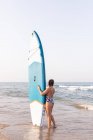 Surfista feminina de pé com placa SUP azul na costa arenosa no verão e olhando para longe — Fotografia de Stock