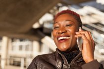 Vista lateral de una hermosa mujer afro negra hablando con su teléfono inteligente mientras sonríe sobre un fondo borroso en un día soleado - foto de stock