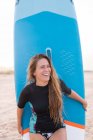 Heureux surfeur féminin debout avec SUP board bleu sur le bord de mer sablonneux en été et regardant loin — Photo de stock