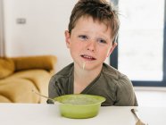 Ragazzo disgustato con la bocca sporca seduto a tavola con ciotola di zuppa di crema durante il pranzo — Foto stock