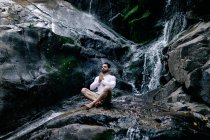 Tranquillo maschio seduto in posa Lotus con le mani di preghiera sulla roccia vicino alla cascata e meditare durante lo yoga con gli occhi chiusi — Foto stock