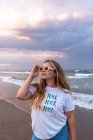 Позитивна молода жінка в модних сонцезахисних окулярах і стильному вбранні, що стоїть на морі проти моря в літній вечір — стокове фото