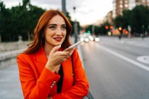 Усміхнена жінка з рудим волоссям і в помаранчевому костюмі записує аудіоповідомлення на мобільний телефон під час спілкування з другом у соціальних мережах і стоячи на міській вулиці — стокове фото