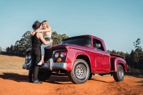 Cool boyfriend embracing smiling girlfriend sitting on hood of red retro pickup parked on sandy road — Stock Photo