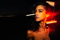 Jeune femme hispanique confiante avec de longs cheveux ondulés et les épaules nues en gardant la main sur la tête et en regardant la caméra tout en se tenant près de la fenêtre en verre avec des reflets de lumière dans l'obscurité la nuit — Photo de stock