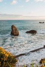 Espectacular paisaje de áspera costa rocosa bañada por espumosas olas marinas a la luz del sol bajo el cielo azul nublado en Liencres Cantabria en España - foto de stock