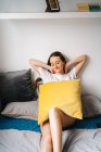 Donna deliziata seduta su un letto morbido con cuscino e godendo il fine settimana con gli occhi chiusi a casa — Foto stock