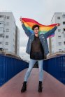 Снизу серьезный гомосексуальный мужчина стоит с радужным флагом в поднятых руках на мосту и смотрит в камеру — стоковое фото