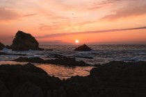 Amazing peaceful scenery of sunset over rippled wavy sea with rocks under colorful cloudy sky in summer evening in Liencres Cantabria Spain — Stock Photo