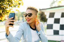 Conteúdo maduro afro-americano feminino em óculos de sol modernos ter vídeo chat no celular no parque em volta iluminado — Fotografia de Stock