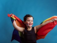 Menina sorridente com bochecha pintada levantando braços com bandeira multicolorida em fundo azul vívido — Fotografia de Stock
