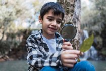 Bambino focalizzato con foglia di pianta verde guardando attraverso lente d'ingrandimento in legno su sfondo sfocato — Foto stock