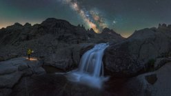 Анонімний мандрівник з факелом, який споглядає водоспад серед грубих скелястих місцевостей під нічним зоряним небом з яскравим світлом Чумацький Шлях — стокове фото