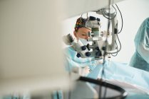 Attentive female doctor in surgical uniform and sterile mask looking through microscope while operating eye of unrecognizable patient in hospital — Stock Photo