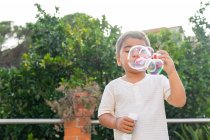Милый маленький мальчик в белой футболке, надувающий мыльные пузыри, стоя в зеленом саду в летний день — стоковое фото