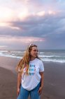 Позитивная молодая женщина в модных солнцезащитных очках и стильном наряде, стоящая на берегу моря в летний вечер — стоковое фото
