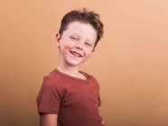 Удовлетворенный ребенок в повседневной одежде с каштановыми волосами, смотрящий в камеру с зубчатой улыбкой и наклоненной головой — стоковое фото