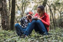 Ethnic girl writing in notepad against brother looking through binoculars while sitting on land in summer woods — Stock Photo
