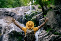 Back view of unrecognizable male hiker standing on boulder and admiring waterfall in forest with open arms — Stock Photo