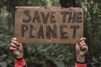 Zugeschnittenes unkenntliches ethnisches Kind zeigt Save The Planet-Titel auf Kartonstück, während es im Wald in die Kamera schaut — Stockfoto