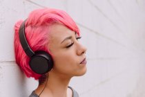 Молода жінка з яскраво-рожевим волоссям слухає музику з навушниками, стоячи біля білої стіни з закритими очима — стокове фото