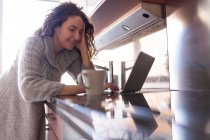 Mulher de negócios com cabelo encaracolado na cozinha tomando uma infusão ao usar seu laptop e trabalhando em casa — Fotografia de Stock